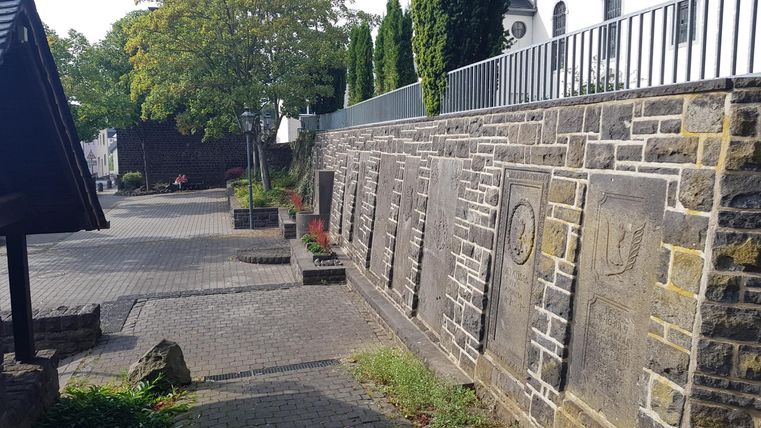 Eine gepflasterte Straße mit Steinen und einer Mauer im Hintergrund. Auf der linken Seite stehen einige Bäume und Pflanzen.