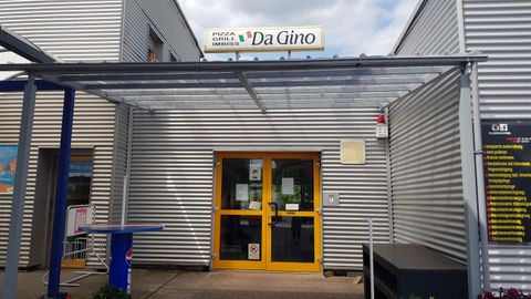 Ein Eingangsbereich zu einem Restaurant mit dem Namen "Da Gino". Die Fassade ist aus grauem Metall mit gelben Türen und einem Überdach.