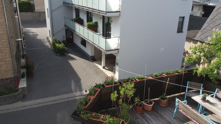 A modern residential building with balconies and plants along the facade. In the foreground, garden plants and a terrace are visible.