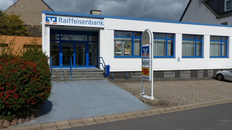 Ein modernes Bankgebäude der Raiffeisenbank mit blauen Fenstern und einer einladenden Eingangstür. Der Gehweg ist gepflastert und es gibt eine Grünanlage vor dem Gebäude.