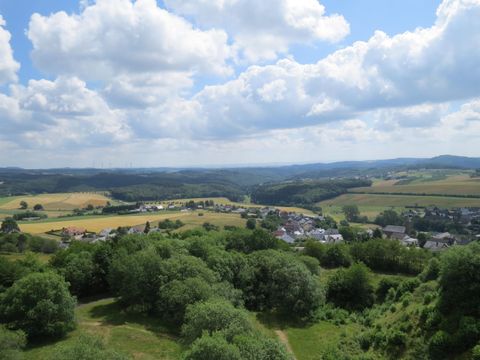 Eine malerische Landschaft mit sanften Hügeln und viel grünem Pflanzenbewuchs. Am Horizont sind kleine Dörfer und ein blauer Himmel mit Wolken zu sehen.