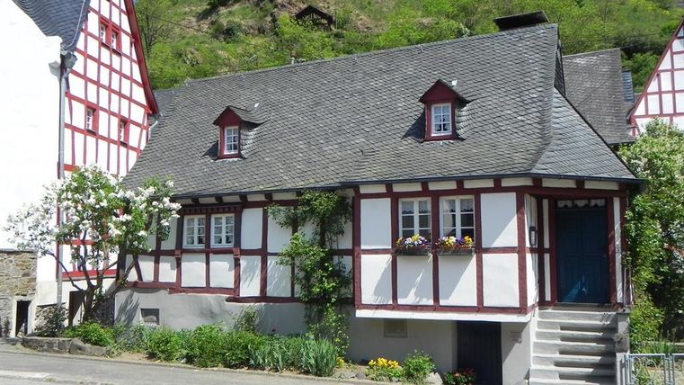 A charming half-timbered house with red and white walls. In the background, green hills and old castle ruins can be seen.