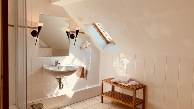 Ein modernes Badezimmer mit einer Dusche, Waschbecken und einem Fenster im Dachbereich. Es gibt eine Holzbank für Handtücher und einen hellen, minimalistischen Stil.