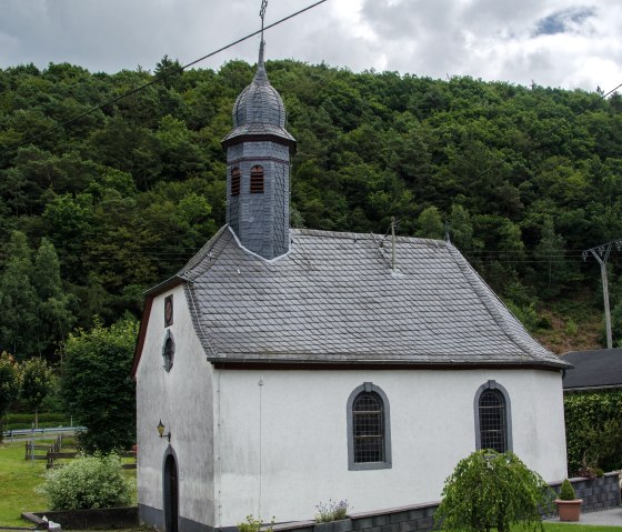 Een kleine kapel met een grijs leistenen dak en toren staat voor een beboste heuvel. De lucht is bewolkt., &copy; Volker Windheuser