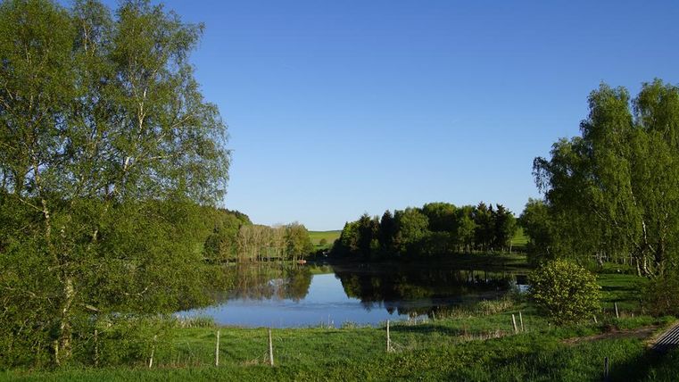 Landschaft mit See, umgeben von Bäumen und Wiesen unter klarem blauem Himmel.