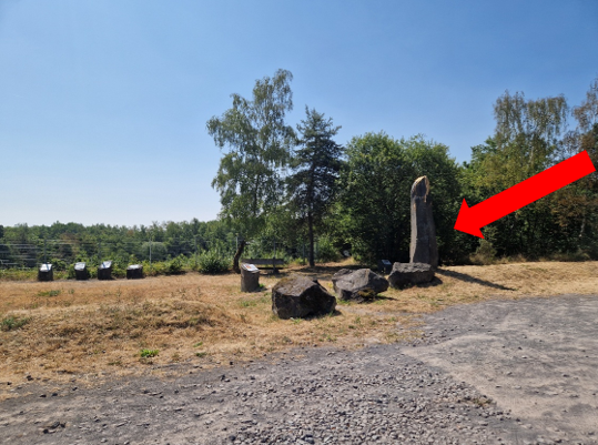 Eine steinerne Statue steht in einer grünen Landschaft mit Bäumen. Im Hintergrund sind weitere Steine auf dem Boden zu sehen.