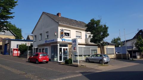 Ein großes Gebäude an einer Straßenecke mit einem Autoparkplatz. Im Vordergrund steht ein rotes Auto und es ist blauer Himmel zu sehen.