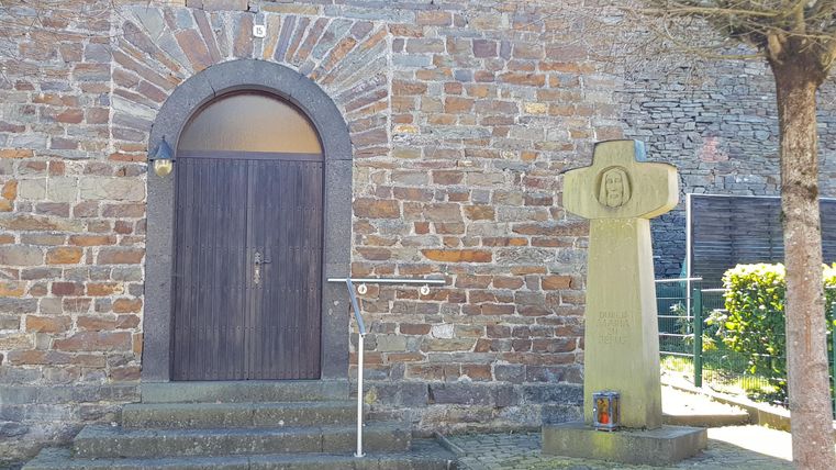 Eine steinerne Kirche mit einer Holztür und einem großen Kreuz daneben. Im Hintergrund sind grüne Büsche zu sehen.