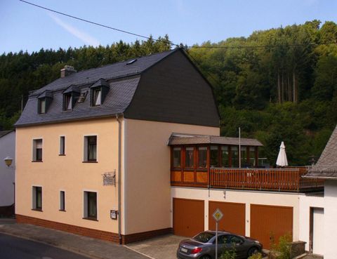 Ein modernes Haus mit einem Holz-Balkon und einer Terrasse. Die Umgebung ist von Bäumen umgeben und die Fassade ist hell gestrichen.