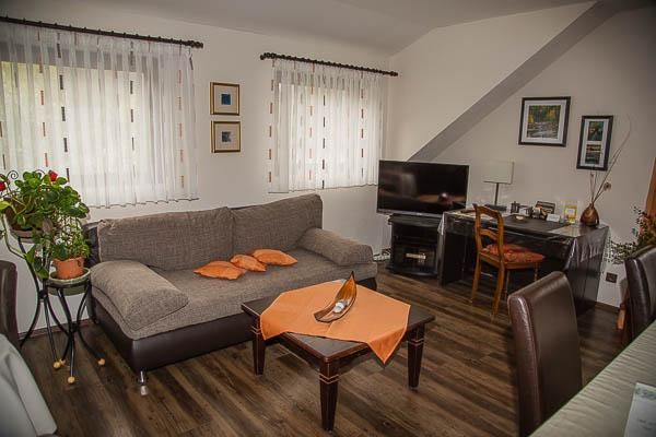 Ein gemütliches Wohnzimmer mit einem grauen Sofa und einem Couchtisch. Am Rand steht ein Schreibtisch mit einem Stuhl und einem Fernseher an der Wand.