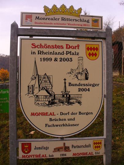 Ein Schild, das das schönste Dorf in Rheinland-Pfalz beschreibt, mit Informationen über Auszeichnungen und Sehenswürdigkeiten. Es zeigt eine Zeichnung von Gebäuden und einer Burg.