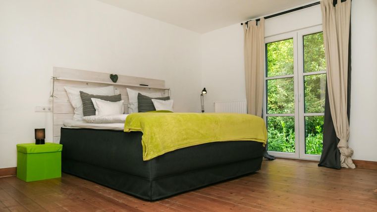 A modern bedroom with a large bed and a bright duvet. The room features large windows overlooking greenery and has a minimalist style.