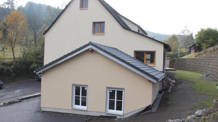 Ein zweigeschossiges Wohnhaus mit einem hellen Außenanstrich. Im Hintergrund sind grüne Hügel und Bäume zu sehen.