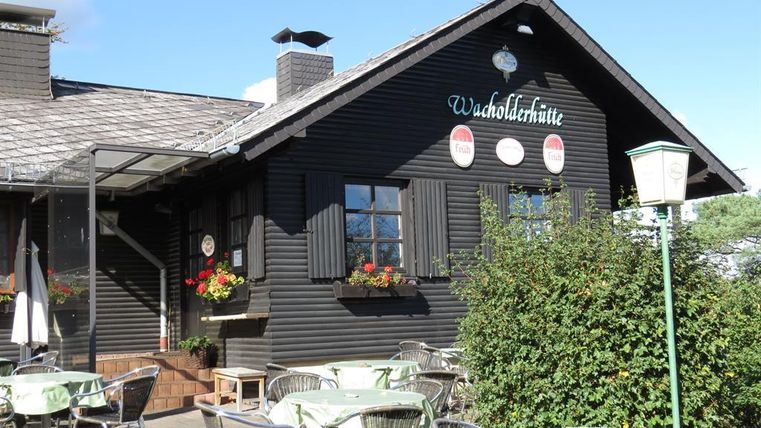 Ein gemütliches, schwarzes Holzgebäude mit Tischen und Stühlen im Freien. Blumen schmücken die Fenster, und ein Schild deutet darauf hin, dass es sich um die „Waldschlütte“ handelt.