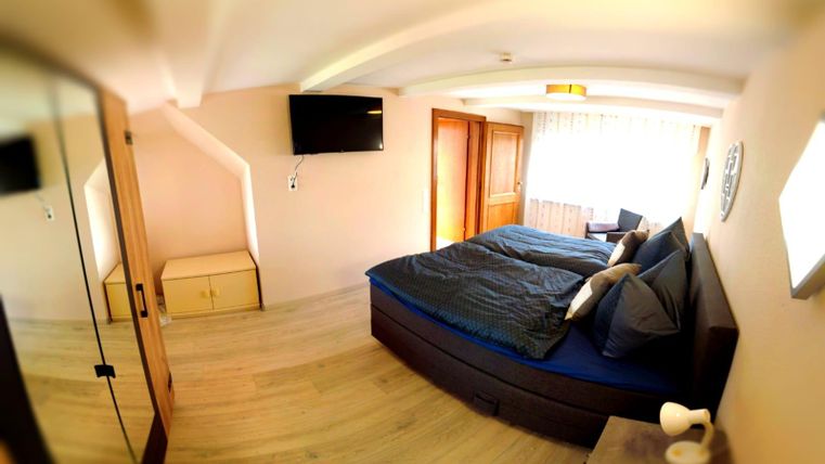 Ein modernes Schlafzimmer mit einem großen Bett und blauer Bettwäsche. An der Wand hängt ein Fernseher und es gibt viel Tageslicht durch das Fenster.