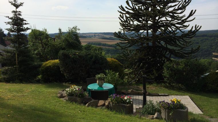 A beautiful garden with a terrace and a table in the middle. In the background, there are trees and a gentle hilly landscape.