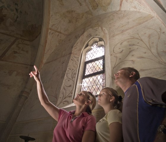 Drei Personen betrachten Fresken in einer Kirche. Eine Frau zeigt auf die Decke. Ein gotisches Fenster mit Gitter ist im Hintergrund sichtbar., &copy; Traumpfade/Kappest