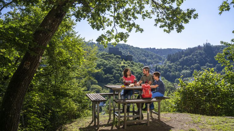 Drei Personen sitzen an einem Picknicktisch im Grünen mit Blick auf eine Burgruine.