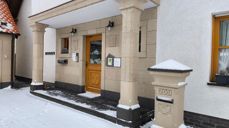 Ein Haus mit einem Eingangsbereich, der von Säulen flankiert ist. Der Boden ist mit Schnee bedeckt und ein Briefkasten steht neben der Tür.