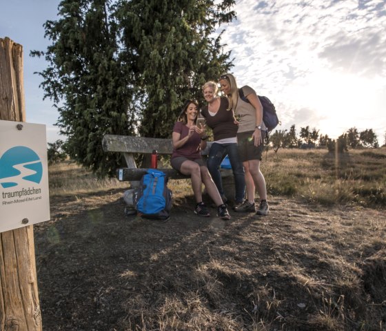 Drei Frauen sitzen auf einer Bank in einer Heidelandschaft, lächelnd und mit Rucksäcken. Ein Schild "traumpfädchen" ist im Vordergrund sichtbar., © Kappest/REMET