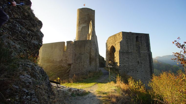 Eine alte Burg steht auf einem Hügel, umgeben von grüner Landschaft. Die Sonne beleuchtet die Ruinen und den alten Turm.