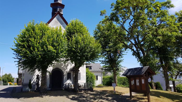 Eine kleine Kirche umgeben von grünen Bäumen und einem klaren Himmel. Im Vordergrund steht ein Informationsschild.