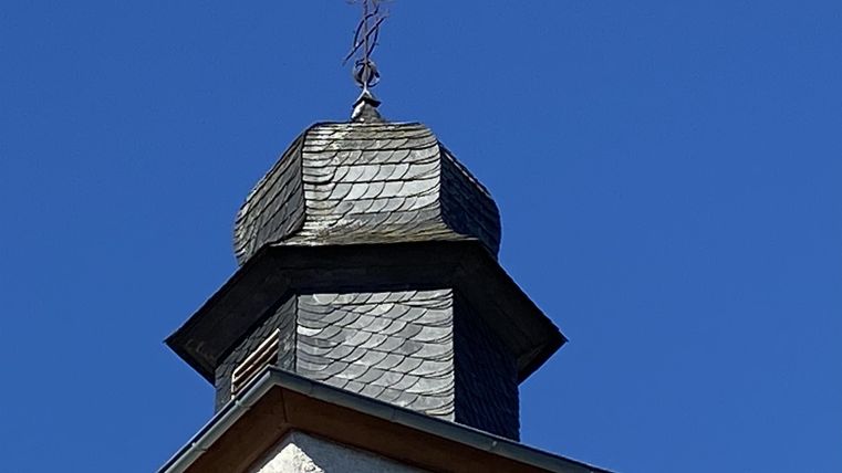 Eine Dachspitze mit einem Schornstein und einer Wetterfahne. Der Himmel ist klar und blau.