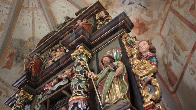 An intricately carved altar with colorful figures. The walls display historical frescoes in a sacred space.