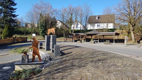 Ein ruhiger Platz mit einem Steinbearbeitungsdenkmal und einem Holzhaus im Hintergrund. Die Umgebung ist von Bäumen umgeben, und die Landschaft wirkt einladend und gepflegt.
