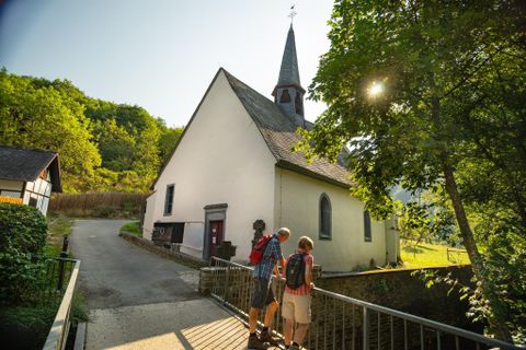 A small church with a tall tower stands in a picturesque landscape. Two hikers are standing on a bridge and enjoying the surroundings.