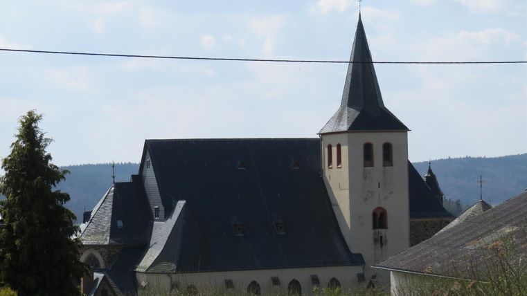 Eine alte Kirche mit einem hohen Turm und einem spitzen Dach. Sie steht in einer ländlichen Umgebung mit Bäumen und Hügeln im Hintergrund.