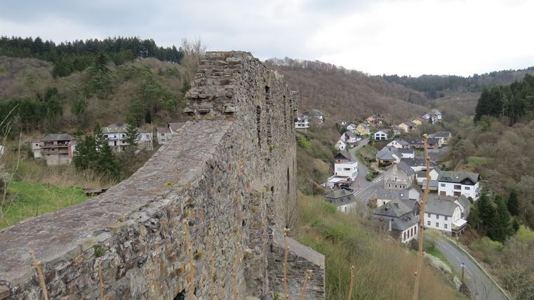 An old castle wall overlooking a small village in the green valley. The sky is cloudy and the landscape is hilly.