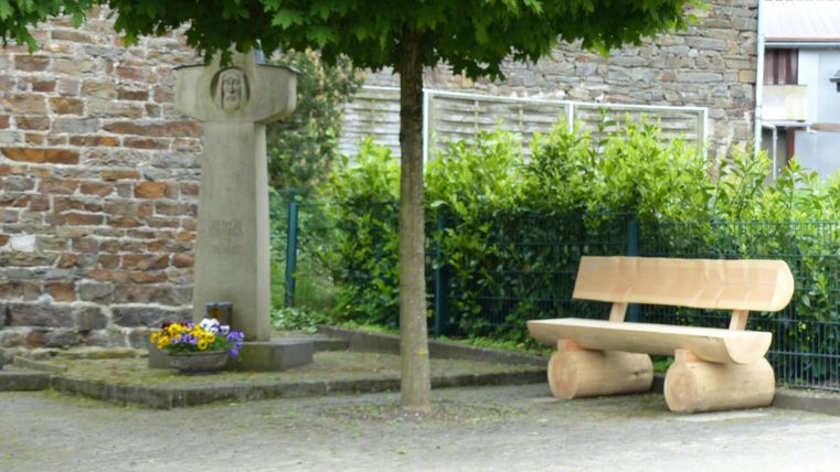 A stone statue with a cross next to a tree-lined square. On the opposite side, there is a large wooden bench and flowers in a planter.