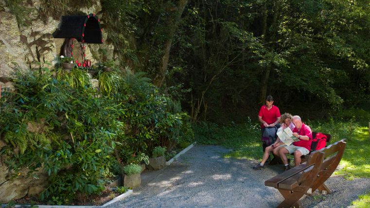 Three people are sitting on a bench in the forest, next to a rock face with a small statue of the Madonna.