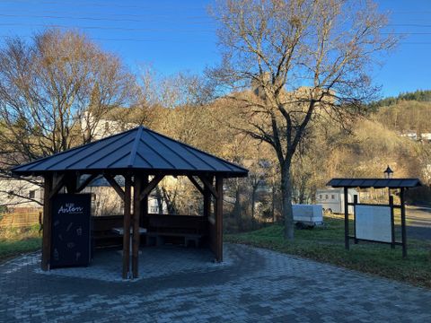 Eine gemütliche Pavillon-Sitzgelegenheit umgeben von Bäumen und Hügeln. Im Hintergrund ist ein Informationstafel zu sehen.