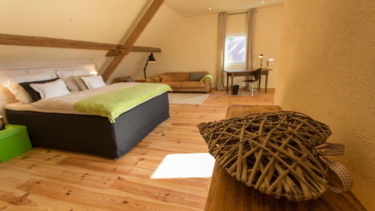 A cozy room with a large bed and warm wooden floors. There is a sitting area and plenty of natural light.