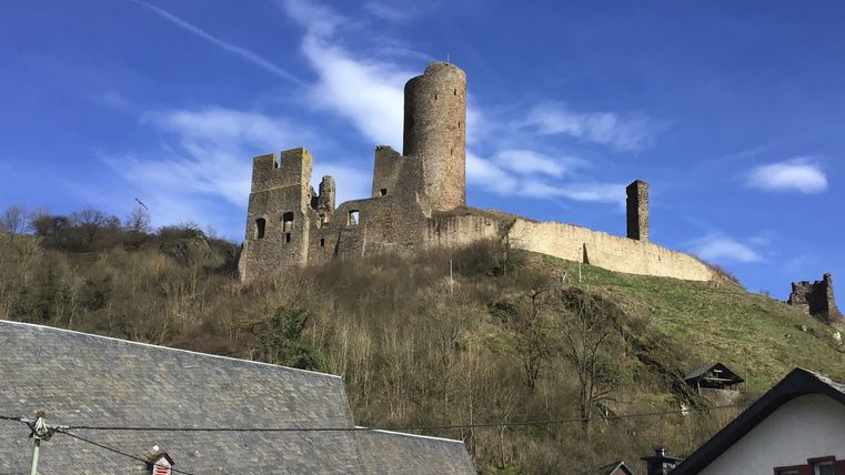 Eine alte Burg thront auf einem Hügel über einem kleinen Dorf. Der Himmel ist klar und blue, und die Umgebung ist grün und bewaldet.