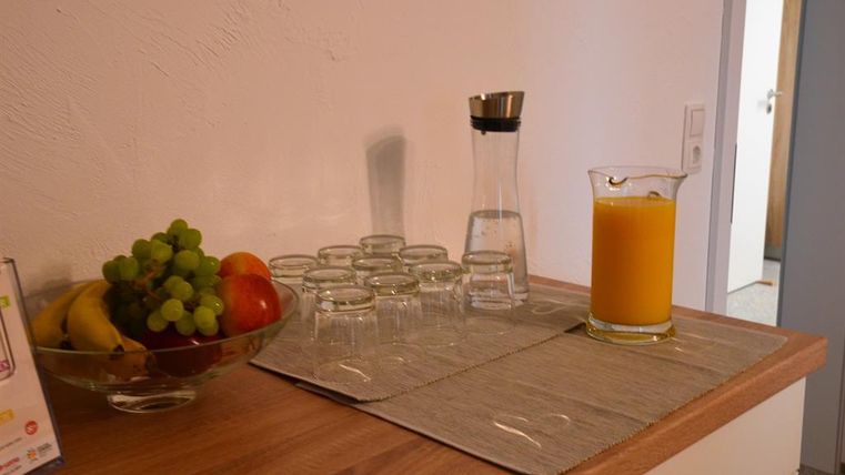 A tabletop with a bowl of fruit, including grapes, apples, and bananas. Next to it are glasses and a carafe of water as well as a glass of orange juice.