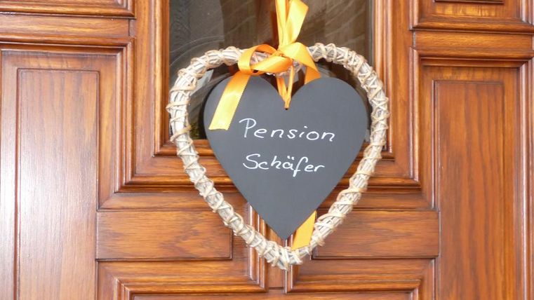 A heart-shaped sign with the inscription "Pension Schäfer" hangs on a wooden door. It is decorated with an orange bow.