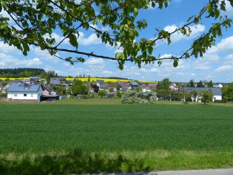 Eine ruhige ländliche Szenerie mit grünen Feldern und kleinen Häusern. Im Hintergrund sind eine schöne Landschaft und ein blauer Himmel mit weißen Wolken zu sehen.