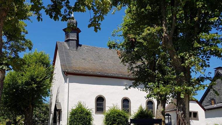 A pretty church with a stone and wooden roof, surrounded by trees. The sky is clear and blue.