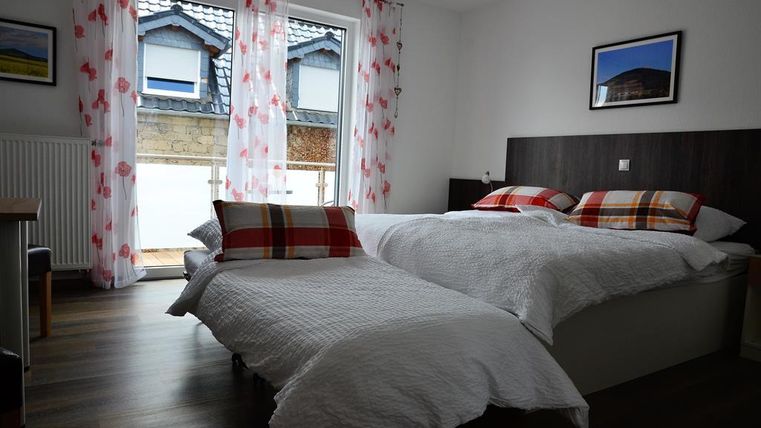 A simple bedroom with two beds and pillows in red and white checkered patterns. Large windows with curtains and a view outside.