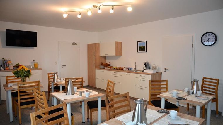 A caretaker room with several tables and chairs. In the background, there is a modern kitchen and a wall with a television and a clock.