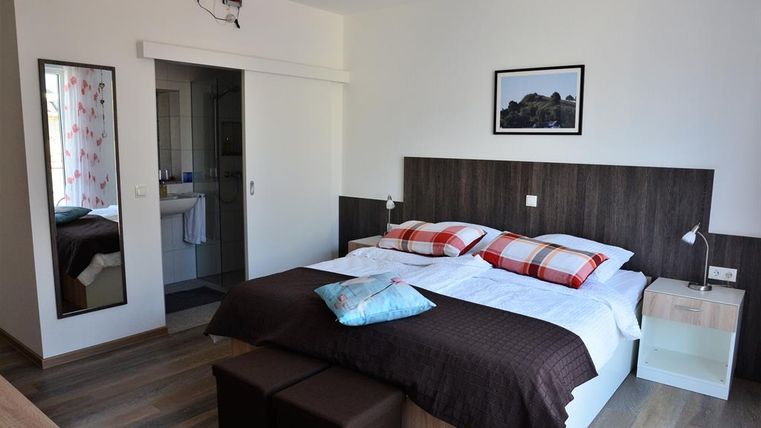 A modern bedroom with a double bed and colorful cushions. The room has a large mirror and access to a bathroom.