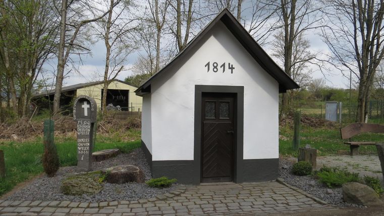 A small, historic building with the year 1814. It is located in a quiet environment, surrounded by trees and stones.