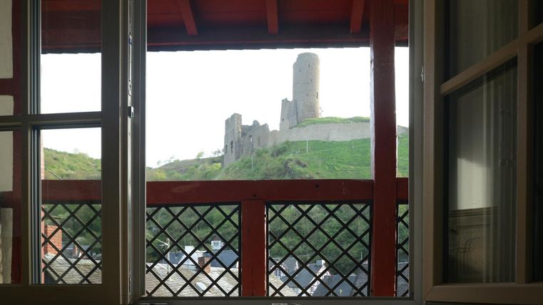 Ein Blick aus einem Fenster auf eine alte Burgruine auf einem Hügel. Die Szenerie ist umgeben von grünen Wiesen und einem klaren Himmel.