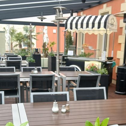A cozy outdoor area of a restaurant with modern tables and chairs. There are plants and umbrellas that create an inviting atmosphere.