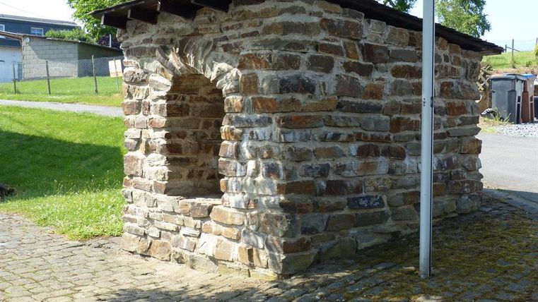 Eine kleine, rustikale Steinhütte mit einem Spitzdach steht auf einem gepflasterten Weg. Im Hintergrund sind Grünflächen und Bäume zu sehen.