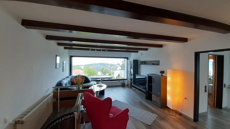 Ein modernes Wohnzimmer mit großen Fenstern, die einen Blick ins Freie bieten. Eine gemütliche Sitzecke ist mit einem roten Sofa und einem stilvollen Tisch eingerichtet.