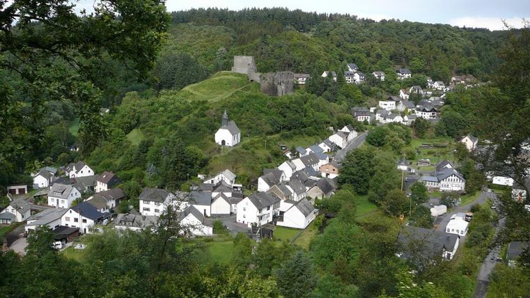 Eine malerische Landschaft mit einem kleinen Dorf, umgeben von Bäumen und Hügeln. Im Hintergrund ist eine alte Burgruine zu sehen.